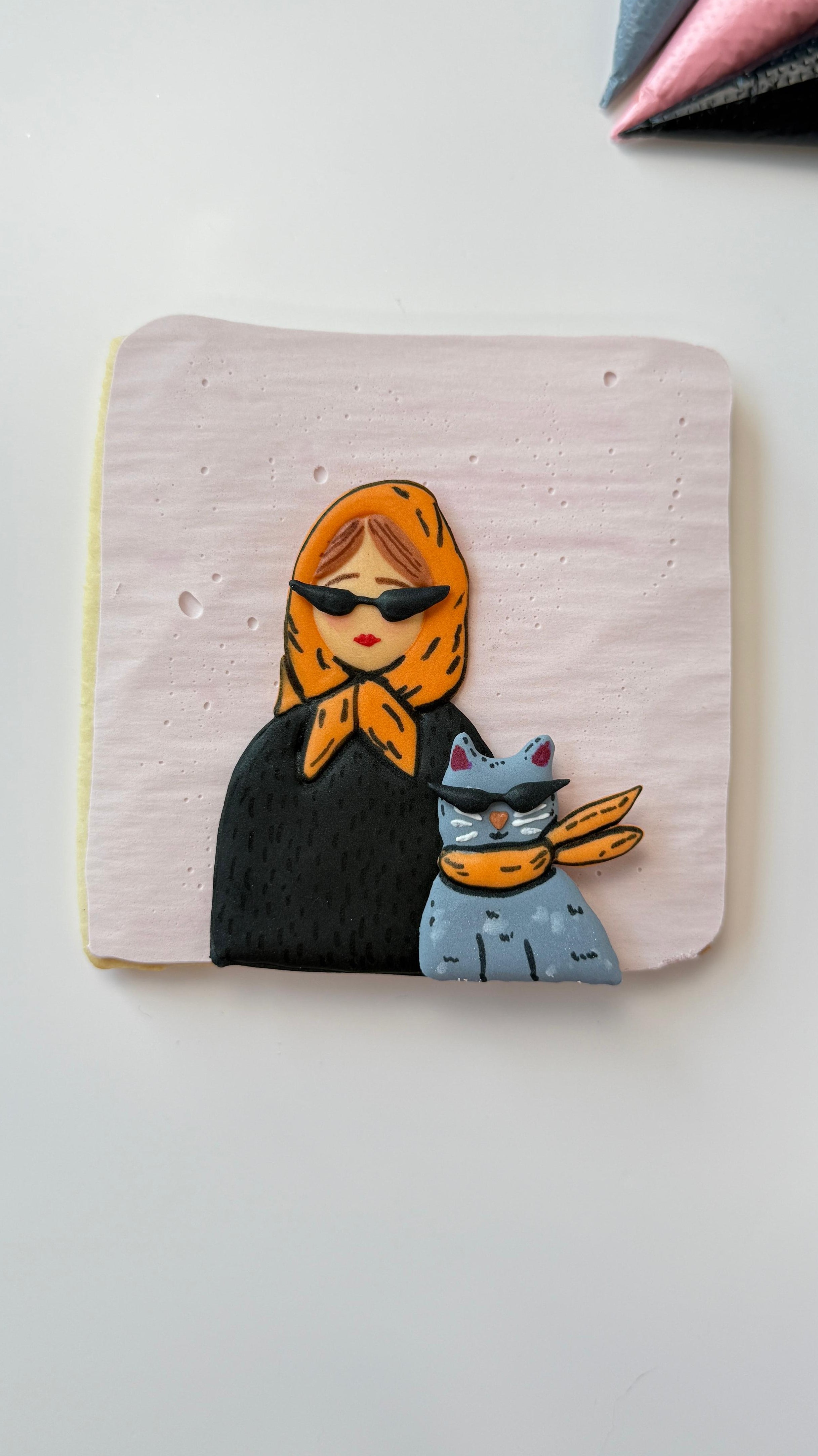 Square decorated cookie with a design of a person wearing an orange headscarf and a cat, on a light pink background.